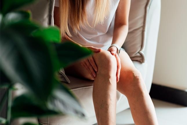 Uma pessoa sentada em um sofá, segurando o joelho, com uma planta em primeiro plano e veias levemente visíveis na perna