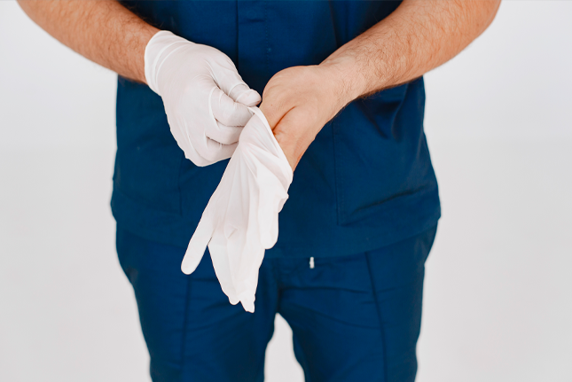 Close-up de um profissional de saúde, vestindo uniforme cirúrgico azul, colocando uma luva de látex branca na mão direita.