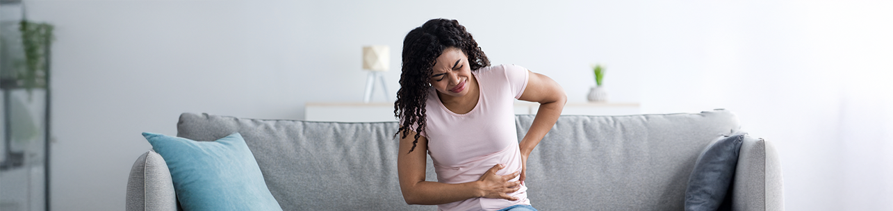 Uma jovem com cabelos encaracolados sentada em um sofá, visivelmente com dor, segurando a parte direita do abdômen e das costas