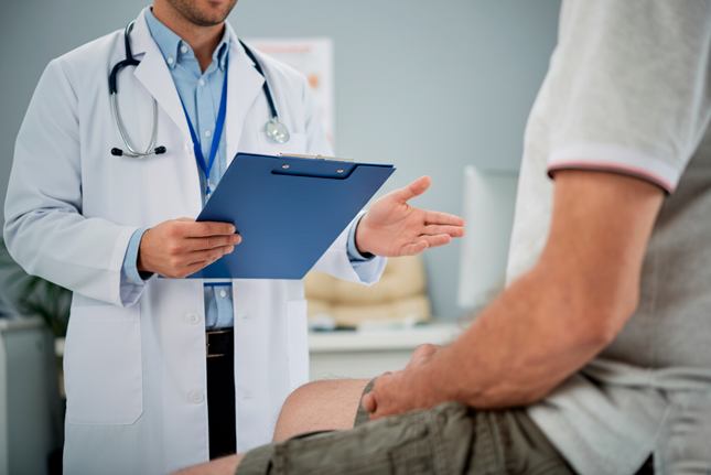 médico em jaleco branco e estetoscópio, segurando uma prancheta azul e gesticulando com a mão para um paciente, que está sentado