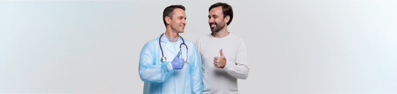 Médico de meia-idade, vestindo bata cirúrgica azul e luvas, e um paciente barbudo sorrindo, ambos olhando um para o outro e fazendo um sinal de 'joinha' (polegar para cima) com as mãos